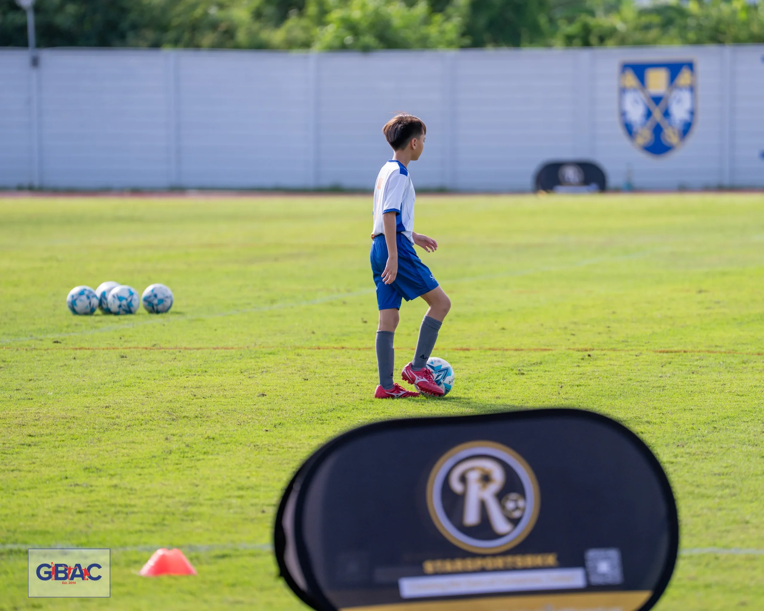 A young football player stands near several footballs during warm-up at a GBAC match